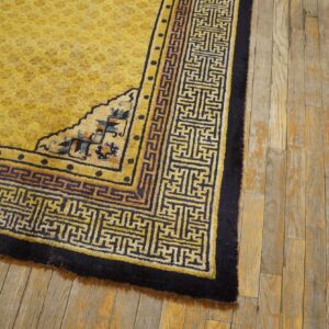 High-pile yellow rug with navy blue fretwork borders rests on distressed wood flooring.