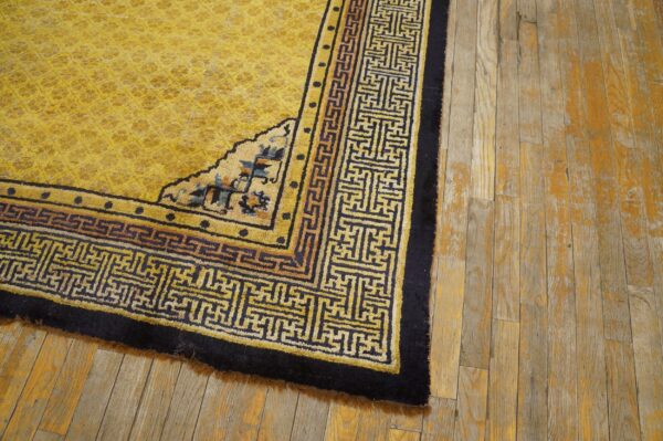 High-pile yellow rug with navy blue fretwork borders rests on distressed wood flooring.