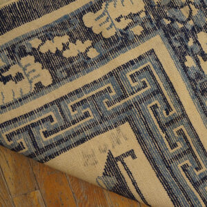 Beige flatweave rug with complex navy and light blue Greek key border lifted above hardwood flooring.