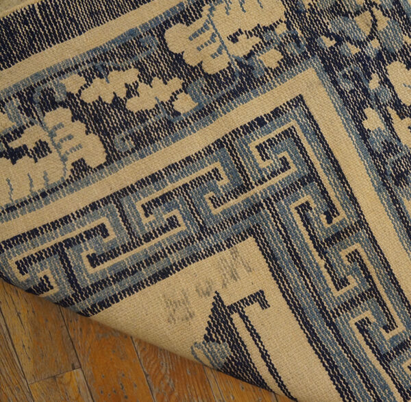 Beige flatweave rug with complex navy and light blue greek key border lifted above hardwood flooring.