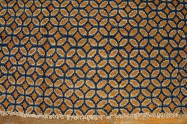 Textured rug with a geometric gold and navy lattice pattern, ending in white fringe on a wood floor.