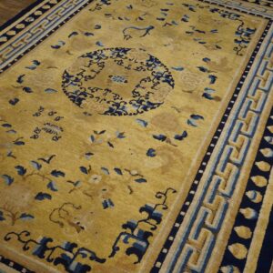 Gold field rug with dark blue stylized floral patterns and a wide geometric fret border on brown wood floors.