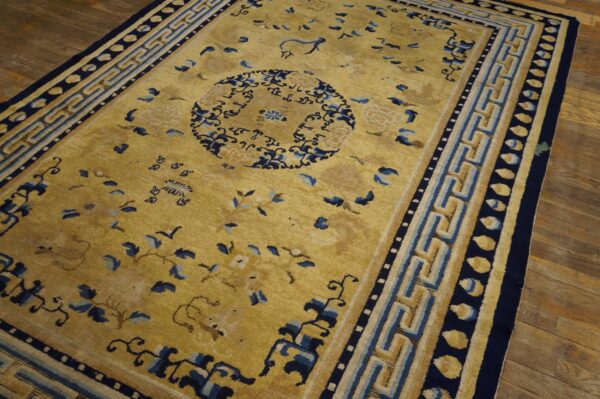 Gold field rug with dark blue stylized floral patterns and a wide geometric fret border on brown wood floors.