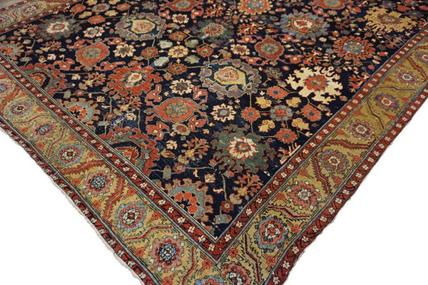 Ornate rug corner featuring a dark blue field, detailed red, gold, and teal floral motifs, and an ochre border.