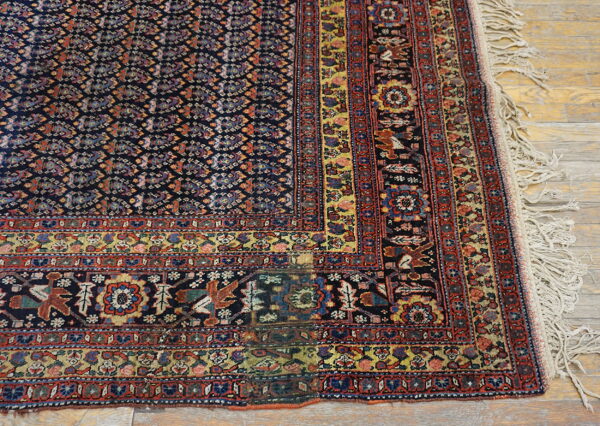 Dark blue rug with complex geometric patterns, red border, and fringe detail rests on hardwood planks.