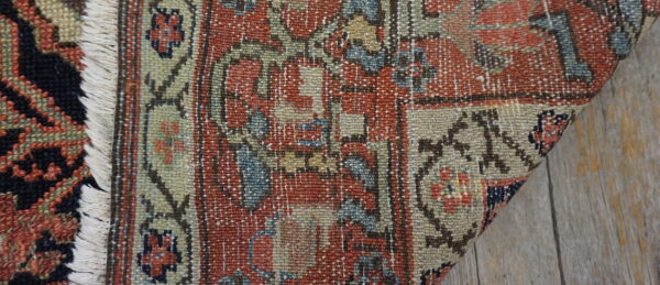 Close-up of a low-pile rug with rust, beige, and blue pattern, white fringe, on plank flooring.