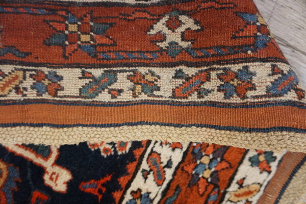 A folded rug displays rust high pile knotting, a cream patterned border, and a flatwoven edge.