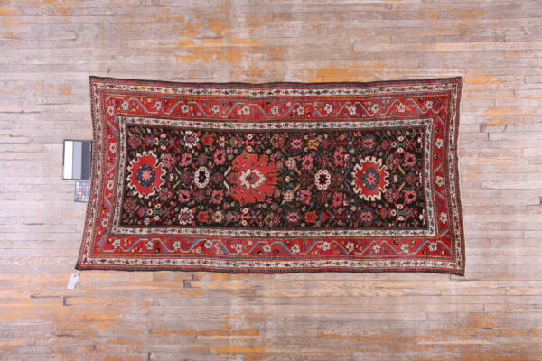 Ornate red and dark floral patterned rug displayed flat on heavily distressed light wood planks.
