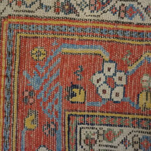 Woven rug detail showing a rust-red field, cream border, and complex blue and yellow patterns.