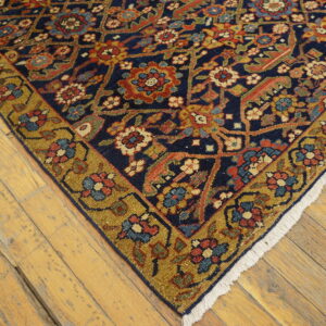 Dark blue floral rug with a gold border and white fringe rests on worn light wood flooring.