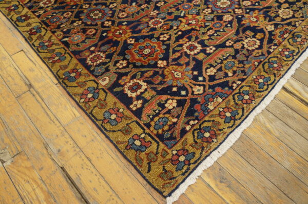Dark blue floral rug with a gold border and white fringe rests on worn light wood flooring.