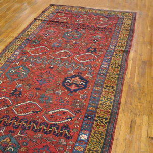 Red rug with detailed geometric patterns in blue and gold lying on light brown hardwood flooring.