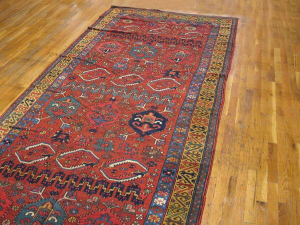 Red rug with detailed geometric patterns in blue and gold lying on light brown hardwood flooring.