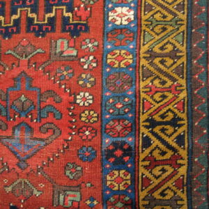 Red geometric rug with stepped motifs and complex gold and blue borders on light wood planks.