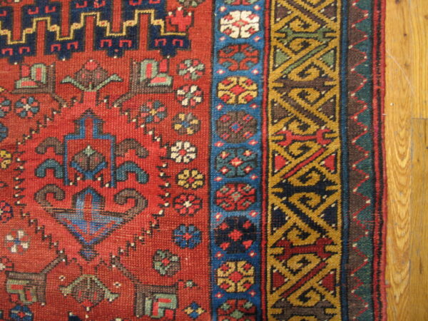 Red geometric rug with stepped motifs and complex gold and blue borders on light wood planks.