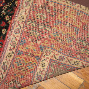 Hand-knotted rug corner lifted, revealing the red, tan, and blue woven backing over wood flooring.