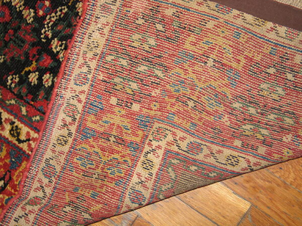 Hand-knotted rug corner lifted, revealing the red, tan, and blue woven backing over wood flooring.