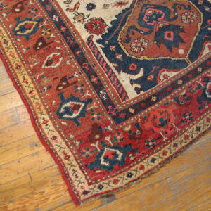 Corner of a traditional rug with rust, navy, and beige patterns on worn, light brown wood planks.