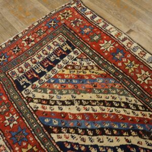 Red, blue, and cream geometric rug with horizontal stripes rests on light, distressed wood floorboards.