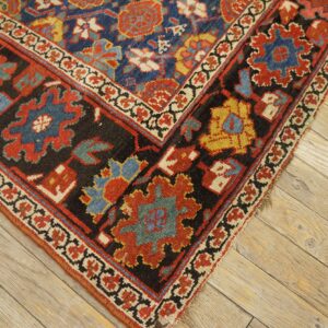 Geometric blue, red, black, and gold patterned rug corner displays low pile on light wood planks.