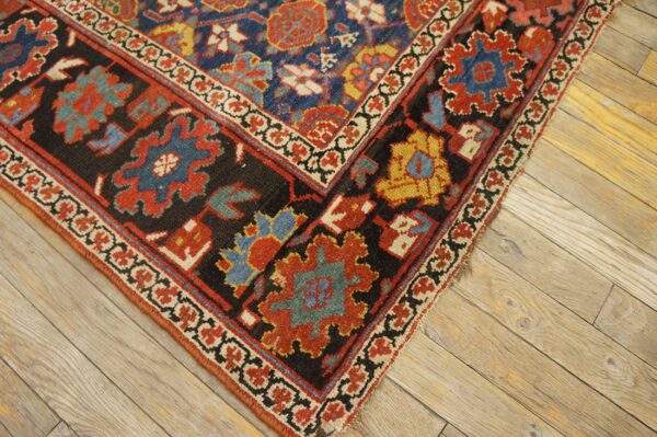 Geometric blue, red, black, and gold patterned rug corner displays low pile on light wood planks.