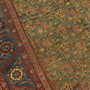 Low-pile rug with a detailed geometric pattern in olive, deep red, and dark blue on light wood flooring.