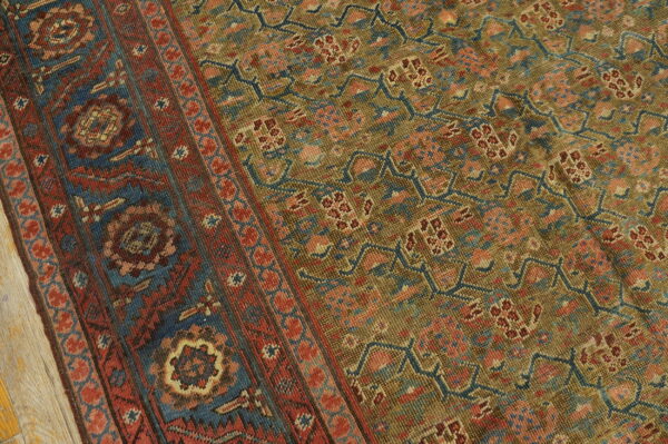 Low-pile rug with a detailed geometric pattern in olive, deep red, and dark blue on light wood flooring.