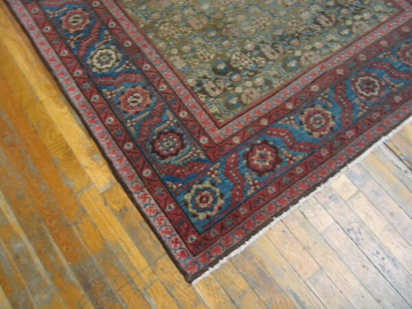 Red and blue patterned border rug with a muted green field lies diagonally on worn wood flooring.