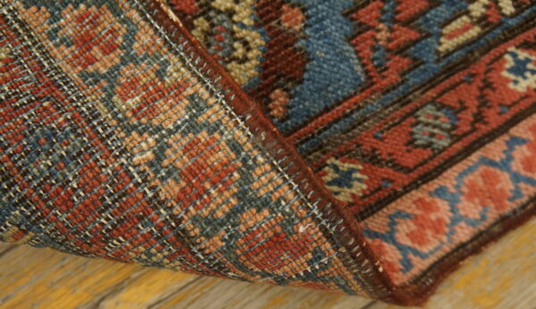 Detail of a rug showing the red and blue pile, woven back, and dark red edge cording on a wood floor.