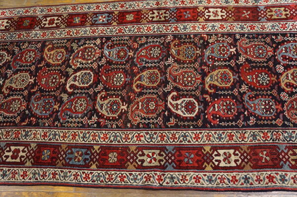 Dark patterned runner rug with repeating red, blue, and tan paisley motifs, bordered by geometrics on wood floor.