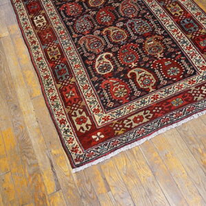 Low-pile rug in red, cream, and dark blue with complex borders sits on distressed light wood floors.