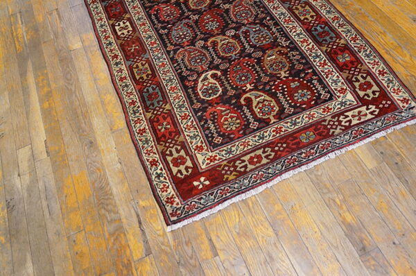 Low-pile rug in red, cream, and dark blue with complex borders sits on distressed light wood floors.