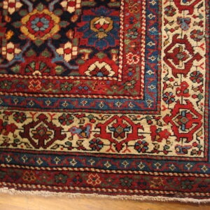 Detailed corner view of a red, navy, and cream traditional rug with intricate borders on light wood flooring.