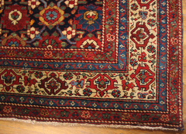 Detailed corner view of a red, navy, and cream traditional rug with intricate borders on light wood flooring.