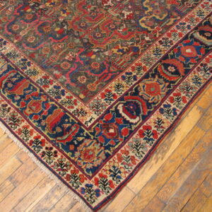 The corner of a low-pile, intricately patterned rug with dark blue and red borders rests on wooden floorboards.
