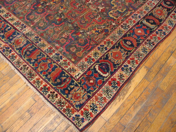 The corner of a low-pile, intricately patterned rug with dark blue and red borders rests on wooden floorboards.