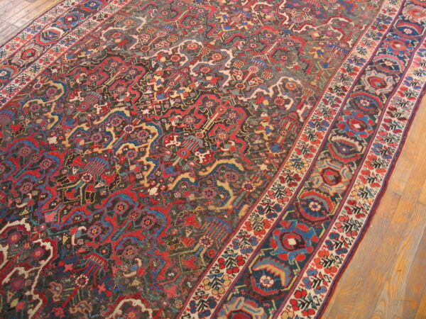 A detailed, low-pile rug with complex red, blue, and dark patterns and borders lies on light wood flooring.