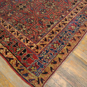 Corner of a low-pile red, blue, and gold traditional rug lying on light angled hardwood flooring.
