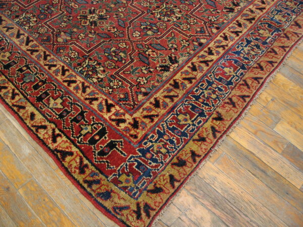 Corner of a low-pile red, blue, and gold traditional rug lying on light angled hardwood flooring.