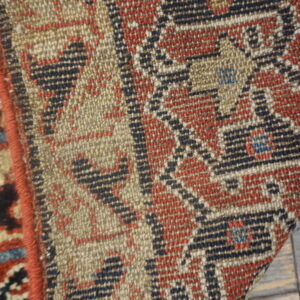 Close-up shows the flat-woven texture and geometric pattern in beige, red, and blue on wood planks.