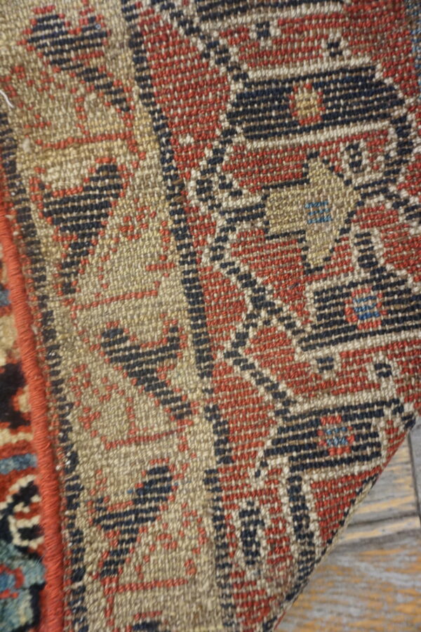 Close-up shows the flat-woven texture and geometric pattern in beige, red, and blue on wood planks.