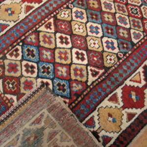 Colorful tribal rug with red, blue, beige, and cream geometric repeating patterns on a wood floor.