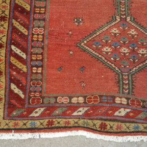 Rust-colored rug with ornate geometric borders, a central diamond motif, fringe, and a concrete background.