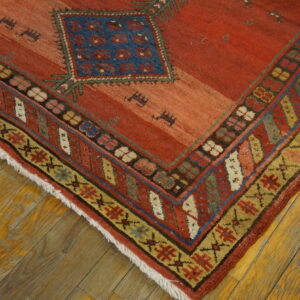 Corner view of a rusty red rug featuring a blue diamond motif and multi-colored geometric borders over wood planks.
