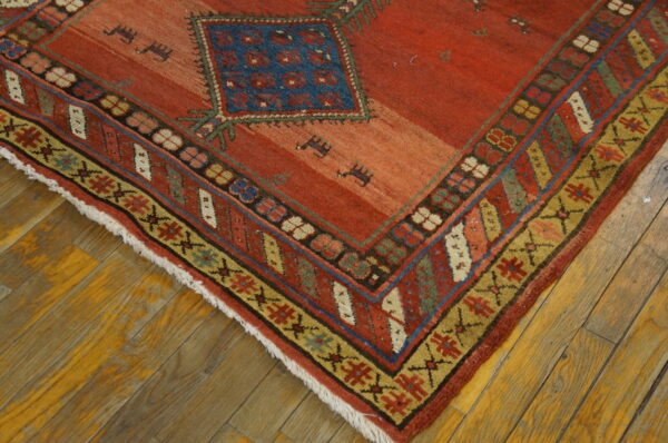 Corner view of a rusty red rug featuring a blue diamond motif and multi-colored geometric borders over wood planks.