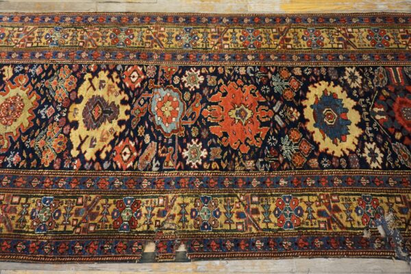Worn runner rug featuring a navy blue field with intricate gold, red, and blue traditional patterns over wood floor.