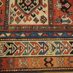 Geometric rug detail featuring dense borders and motifs in terracotta, navy, tan, and light blue resting on a wood floor.