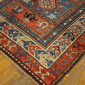 Red and blue geometric rug corner with detailed borders rests on warm wood plank flooring.