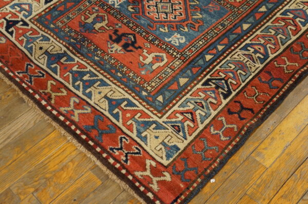 Red and blue geometric rug corner with detailed borders rests on warm wood plank flooring.