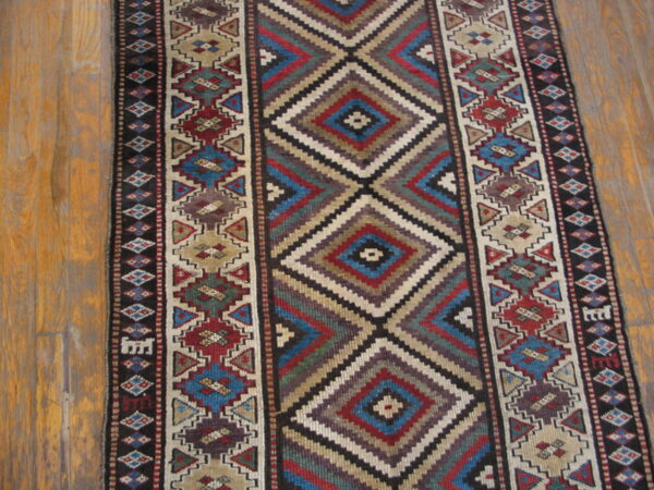 Geometric runner rug in blue, red, tan, and dark tones features stacked diamonds on a wood floor.